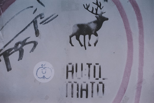 A stencil graffiti of a deer with antlers is sprayed onto a wall, accompanied by layered text reading 'AUTO MATO', a circular sticker with a simplistic drawing of a smiling apple is also visible, and abstract brush-like strokes are on the left side. The overall style appears urban and gritty.