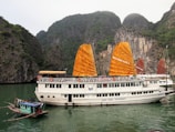 A large white tourist boat with prominent orange sails is anchored on a tranquil green body of water. In the foreground, a smaller boat with a colorful roof floats nearby. The backdrop features towering rock formations covered in lush greenery, creating a picturesque and serene setting.