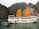 A large white tourist boat with prominent orange sails is anchored on a tranquil green body of water. In the foreground, a smaller boat with a colorful roof floats nearby. The backdrop features towering rock formations covered in lush greenery, creating a picturesque and serene setting.
