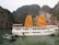 A large white tourist boat with prominent orange sails is anchored on a tranquil green body of water. In the foreground, a smaller boat with a colorful roof floats nearby. The backdrop features towering rock formations covered in lush greenery, creating a picturesque and serene setting.