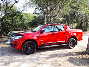 A bright red pickup truck is parked on a dirt surface surrounded by lush green trees, creating a natural setting. The truck has shiny black wheels and the sunlight casts shadows on its side.