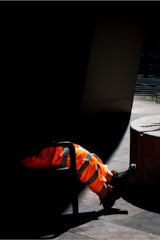 A pair of legs in bright orange reflective work pants and black work boots are partially illuminated by sunlight. The individual is sitting on a chair, with most of their body obscured by shadow, emphasizing the vibrant color of the clothing against the dark background. The setting appears to be an outdoor area with hard flooring and concrete structures.