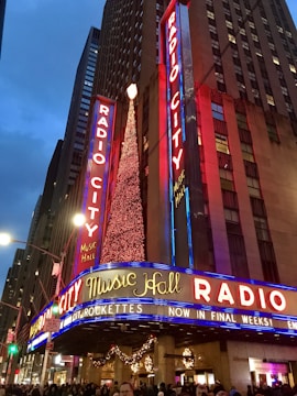 Bright neon lights illuminate the exterior of the Radio City Music Hall. Tall buildings frame the glowing signage, while a large, decorated Christmas tree adds a festive touch. Crowds of people gather at the entrance, creating a lively atmosphere.