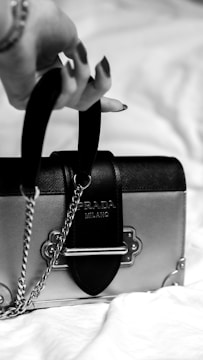 A black and white photograph showcasing a hand holding the handle of a small, elegant handbag with metal accents. The focus is on the intricate details of the bag, which features a plaque with the brand name and a chain strap. The background is softly blurred.