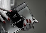 A hand with red polished nails holds a small luxury handbag with the label 'Prada Milano'. The bag features a metallic look with a black textured flap and metal accents. The setting has a subdued and elegant atmosphere.