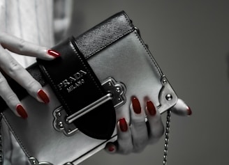 A hand with red polished nails holds a small luxury handbag with the label 'Prada Milano'. The bag features a metallic look with a black textured flap and metal accents. The setting has a subdued and elegant atmosphere.