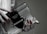 A hand with red polished nails holds a small luxury handbag with the label 'Prada Milano'. The bag features a metallic look with a black textured flap and metal accents. The setting has a subdued and elegant atmosphere.