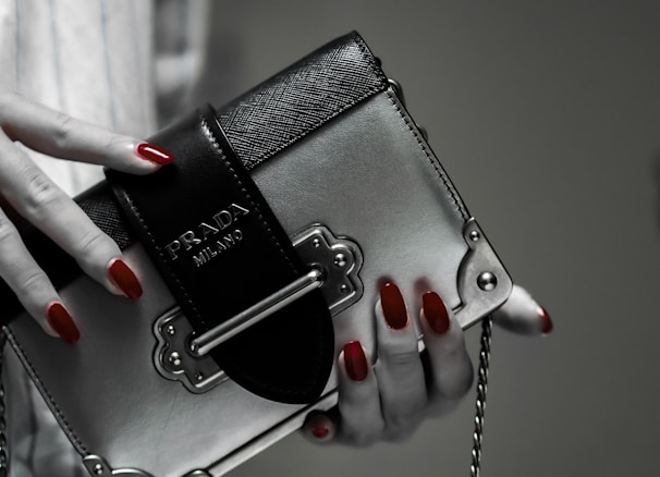 A hand with red polished nails holds a small luxury handbag with the label 'Prada Milano'. The bag features a metallic look with a black textured flap and metal accents. The setting has a subdued and elegant atmosphere.