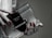 A hand with red polished nails holds a small luxury handbag with the label 'Prada Milano'. The bag features a metallic look with a black textured flap and metal accents. The setting has a subdued and elegant atmosphere.
