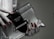 A hand with red polished nails holds a small luxury handbag with the label 'Prada Milano'. The bag features a metallic look with a black textured flap and metal accents. The setting has a subdued and elegant atmosphere.