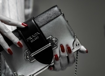 A hand with red polished nails holds a small luxury handbag with the label 'Prada Milano'. The bag features a metallic look with a black textured flap and metal accents. The setting has a subdued and elegant atmosphere.