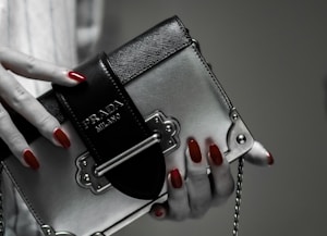 A hand with red polished nails holds a small luxury handbag with the label 'Prada Milano'. The bag features a metallic look with a black textured flap and metal accents. The setting has a subdued and elegant atmosphere.