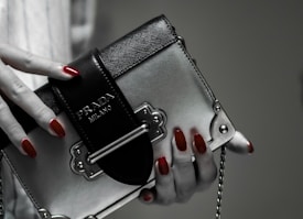 A hand with red polished nails holds a small luxury handbag with the label 'Prada Milano'. The bag features a metallic look with a black textured flap and metal accents. The setting has a subdued and elegant atmosphere.