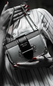 A person holds a small, elegant Prada handbag with a metallic and black design. The bag features a distinct clasp and chain handle. The person's nails are painted red, and they are wearing a striped shirt.