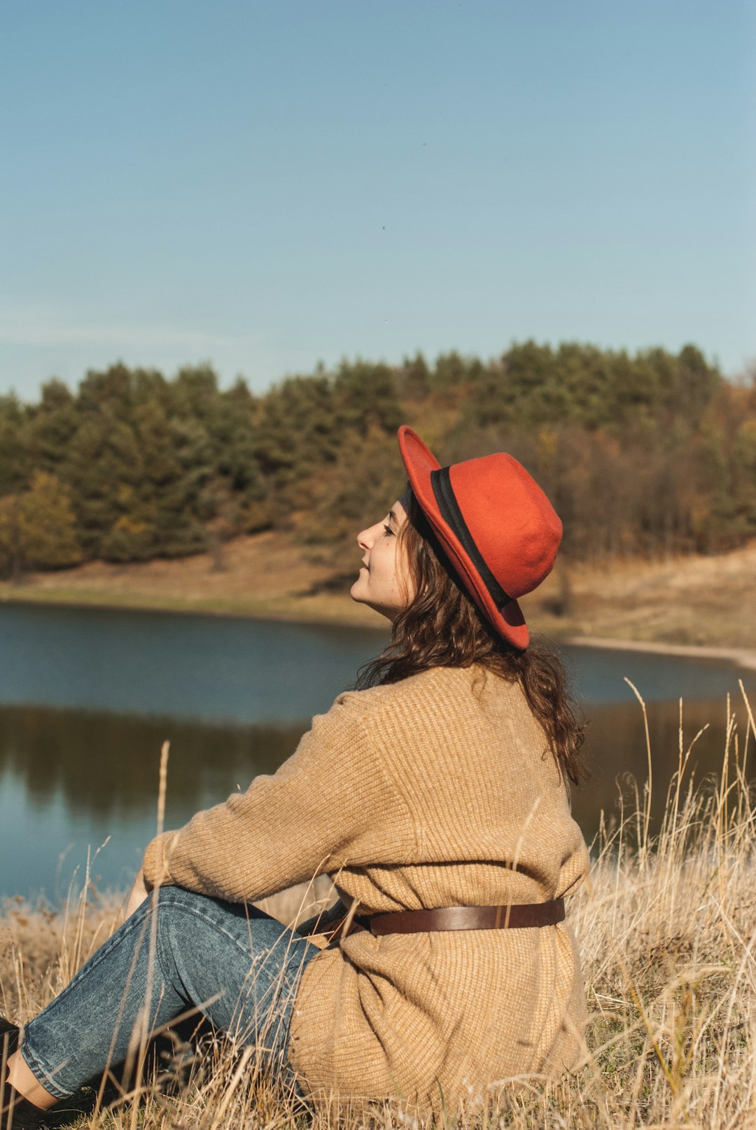 woman in brown sweater and red hat standing on grass field near lake during daytime