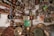 A cluttered room filled with an eclectic mix of vintage and antique items. The scene includes old televisions, copper pots, plates, wall art, and various decorative items. The shelves are crowded with an assortment of objects, including ceramics, dolls, and metallic artifacts. The walls are adorned with colorful plates and framed artworks.