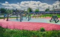 A colorful playground featuring various climbing structures and swings, set in a park with lush green grass and trees in the background. The textured ground features areas of bright red, blue, and green, adding a vibrant feel.