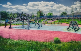 A colorful playground featuring various climbing structures and swings, set in a park with lush green grass and trees in the background. The textured ground features areas of bright red, blue, and green, adding a vibrant feel.