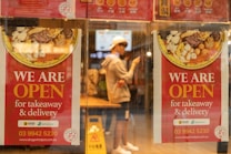 Two red posters on a glass window advertise that a restaurant is open for takeaway and delivery. They feature images of a bowl with various ingredients. A person in casual clothing and a white cap is visible through the window, standing inside the restaurant and using a smartphone.
