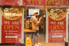 Two red posters on a glass window advertise that a restaurant is open for takeaway and delivery. They feature images of a bowl with various ingredients. A person in casual clothing and a white cap is visible through the window, standing inside the restaurant and using a smartphone.