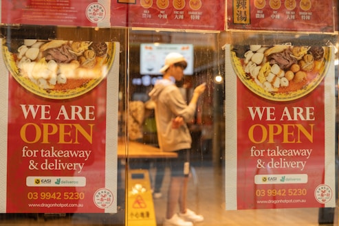 Two red posters on a glass window advertise that a restaurant is open for takeaway and delivery. They feature images of a bowl with various ingredients. A person in casual clothing and a white cap is visible through the window, standing inside the restaurant and using a smartphone.