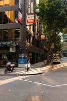 A city street lined with modern buildings features parked vehicles, including a motorcycle and a car. A man stands near a curbside advertisement promoting office spaces for sale. The scene is illuminated by soft sunlight filtering through the trees, casting shadows on the sidewalk and road. The surrounding buildings have large windows reflecting the urban environment.