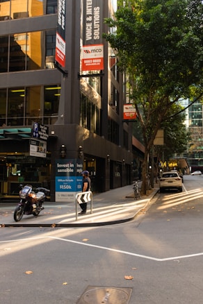 A city street lined with modern buildings features parked vehicles, including a motorcycle and a car. A man stands near a curbside advertisement promoting office spaces for sale. The scene is illuminated by soft sunlight filtering through the trees, casting shadows on the sidewalk and road. The surrounding buildings have large windows reflecting the urban environment.