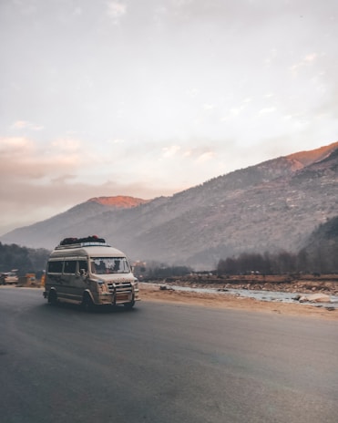 A van travels on a desolate road beside a river with a backdrop of rolling hills under an overcast sky. The morning or evening light casts a warm glow on the peaks of the hills.