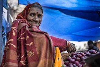 An elderly woman is sitting at a market stall under a blue canopy. She is wearing a traditional red and brown sari with floral patterns. A basket of red onions is placed in front of her. The scene conveys a sense of cultural richness and everyday life.