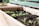 Wooden planter boxes filled with dark soil and small, green seedlings. Labels are attached to sticks, marking different plant sections. The background includes a blurred view of a green bench.