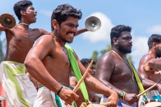 A group of men energetically participate in a traditional festival, playing percussion instruments and cymbals. They are dressed in traditional attire, with vibrant patterns and fabrics draped over their shoulders. The outdoor setting is bright, with a clear blue sky in the background, contributing to the lively atmosphere.