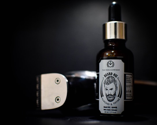 A bottle of beard oil with a silver dropper cap is displayed prominently. The label features an illustration of a bearded man and branding details. Beside it, a metallic clipper head is partially visible against a dark background.