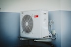 A white outdoor air conditioning unit mounted on a wall with a large fan and electrical connections. The unit has a red label and is surrounded by a light grey and white color scheme on the building's wall.