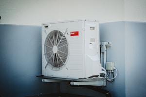 A white outdoor air conditioning unit mounted on a wall with a large fan and electrical connections. The unit has a red label and is surrounded by a light grey and white color scheme on the building's wall.
