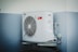 A white outdoor air conditioning unit mounted on a wall with a large fan and electrical connections. The unit has a red label and is surrounded by a light grey and white color scheme on the building's wall.