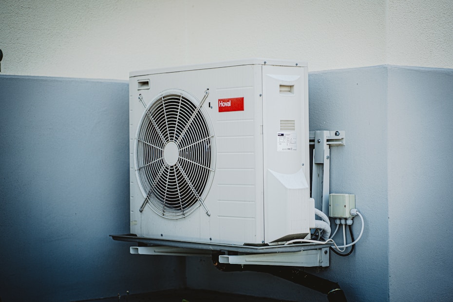A white outdoor air conditioning unit mounted on a wall with a large fan and electrical connections. The unit has a red label and is surrounded by a light grey and white color scheme on the building's wall.