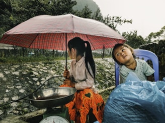 Two children are seated on a roadside under a red checkered umbrella. The child on the left is holding the umbrella, while the child on the right is leaning back slightly. They are surrounded by nature, with a stone wall in the background and greenery. There is a blue sack and a weighing scale visible near them.