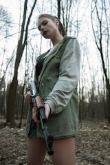 A person stands in a forest holding a rifle, wearing a green jacket with a grey sleeve and looking directly at the camera. The background consists of tall, leafless trees.
