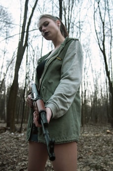 A person stands in a forest holding a rifle, wearing a green jacket with a grey sleeve and looking directly at the camera. The background consists of tall, leafless trees.