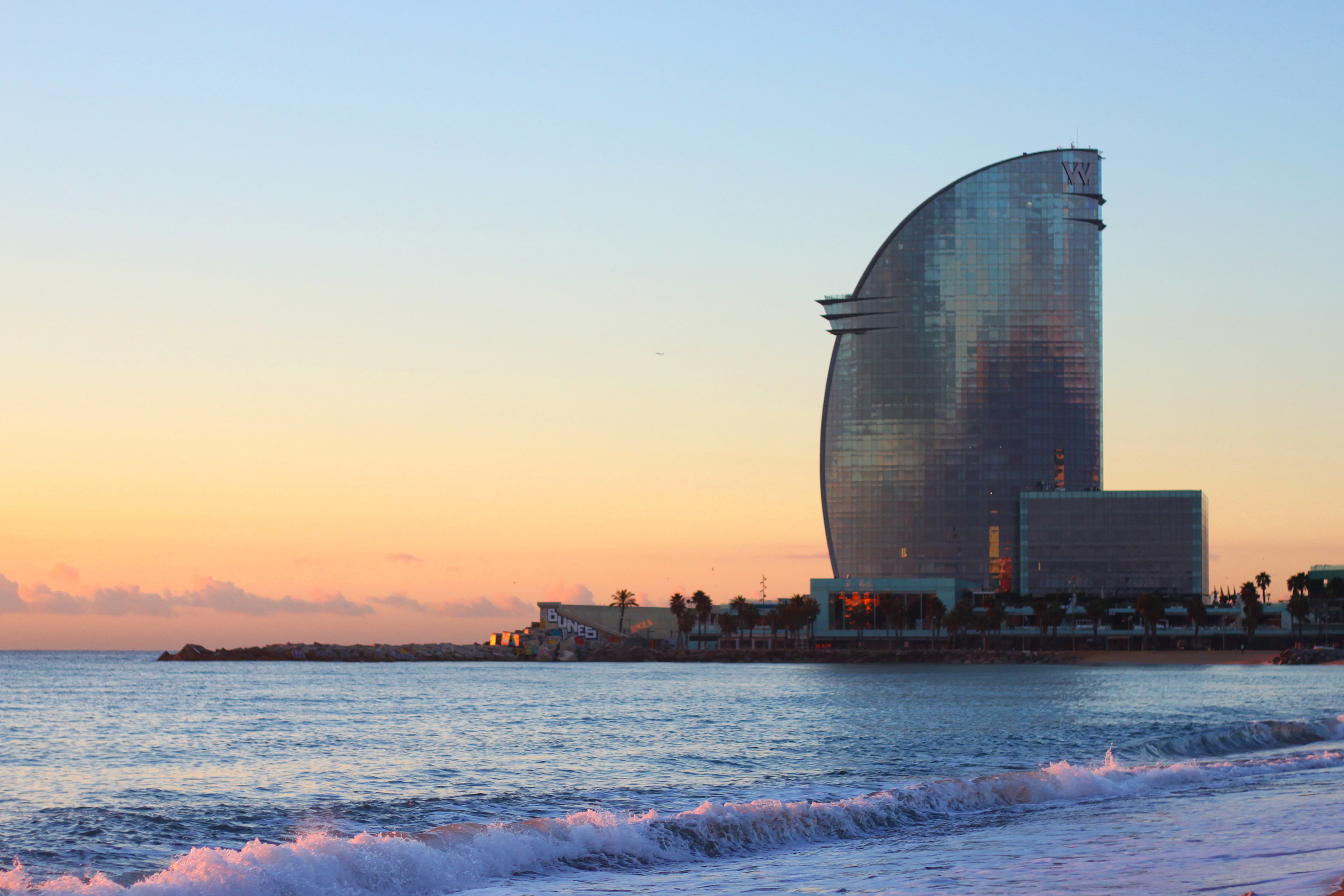 The sail-shaped glass tower of the W hotel standing over Barceloneta beach at first light, small waves breaking on the sand under a peach and blue sky.