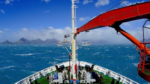 A ship's deck facing a vast and turbulent ocean with distant misty mountains on the horizon. A large red crane is prominently featured on the ship, extending over the blue-green sea. The sky is partly cloudy, suggesting windy conditions as evidenced by the rough sea.