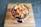 A colorful serving of fresh salsa in a blue-rimmed glass bowl placed on a wooden surface. The salsa contains chopped tomatoes, onions, green peppers, and possibly cucumbers, garnished with two tortilla chips.