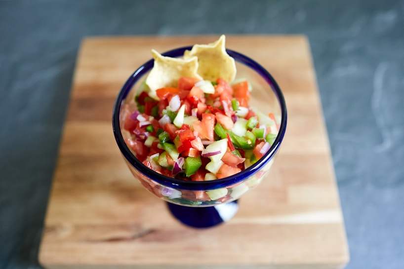 A colorful serving of fresh salsa in a blue-rimmed glass bowl placed on a wooden surface. The salsa contains chopped tomatoes, onions, green peppers, and possibly cucumbers, garnished with two tortilla chips.