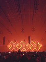 A large indoor concert venue with a crowd of people facing a stage. The stage is illuminated with an array of bright lights in geometric patterns, featuring a prominent 'X' shape in vibrant red and orange tones. The ceiling is lined with numerous lighting fixtures, contributing to the dynamic ambiance of the scene.