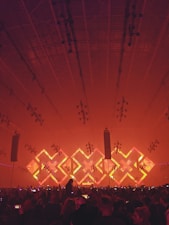 A large indoor concert venue with a crowd of people facing a stage. The stage is illuminated with an array of bright lights in geometric patterns, featuring a prominent 'X' shape in vibrant red and orange tones. The ceiling is lined with numerous lighting fixtures, contributing to the dynamic ambiance of the scene.
