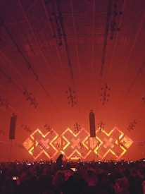 A large indoor concert venue with a crowd of people facing a stage. The stage is illuminated with an array of bright lights in geometric patterns, featuring a prominent 'X' shape in vibrant red and orange tones. The ceiling is lined with numerous lighting fixtures, contributing to the dynamic ambiance of the scene.