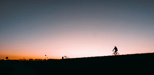 Sunset silhouette of a lone cyclist atop a hill, overlooking rolling gravel trails and distant mountains.