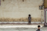 A young child is standing against an old, weathered wall with visible signs of wear and peeling paint. The child appears to be barefoot and is looking downwards, creating a sense of solitude or contemplation. In the foreground, another child, dressed in patterned clothing, walks by, partially out of focus. The ground is a mix of tiled sidewalk and rough gravel.