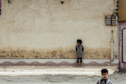 A young child is standing against an old, weathered wall with visible signs of wear and peeling paint. The child appears to be barefoot and is looking downwards, creating a sense of solitude or contemplation. In the foreground, another child, dressed in patterned clothing, walks by, partially out of focus. The ground is a mix of tiled sidewalk and rough gravel.