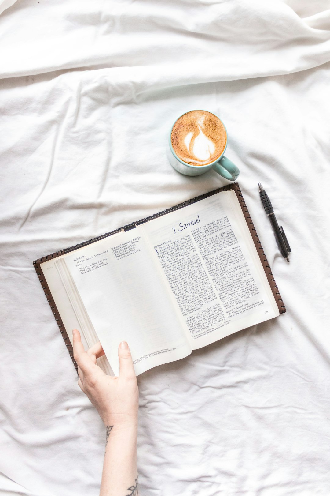 person holding white book page beside black click pen and white ceramic mug on white textile